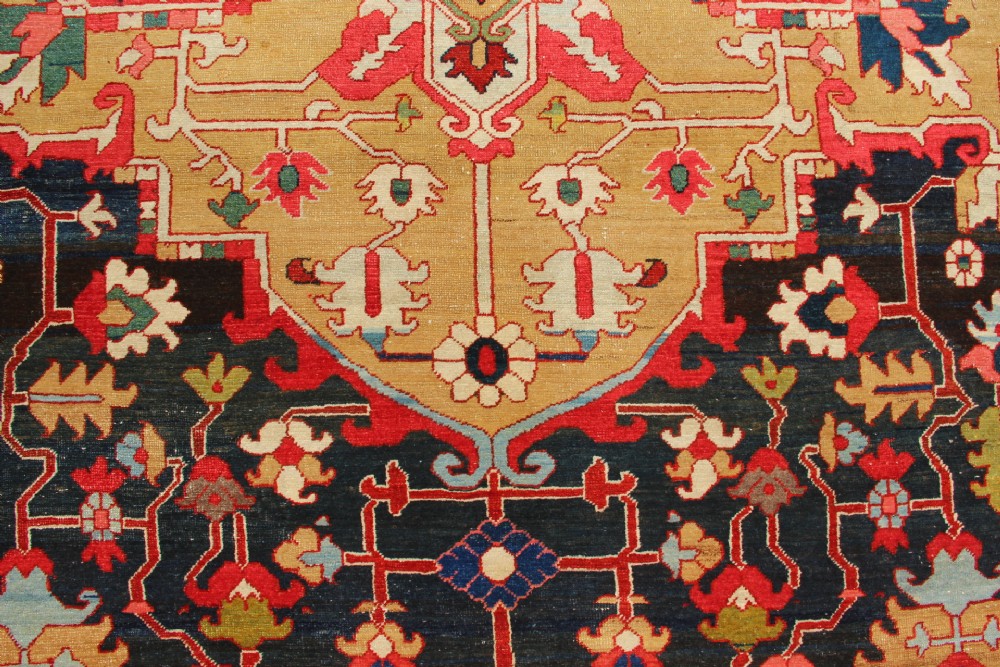 A large hand-knotted Persian Serapi carpet featuring a central geometric medallion in ochre and navy blue, surrounded by a navy field and wide terracotta red border. View V