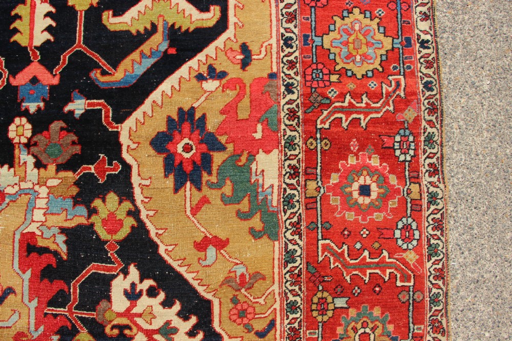 A large hand-knotted Persian Serapi carpet featuring a central geometric medallion in ochre and navy blue, surrounded by a navy field and wide terracotta red border. View VII