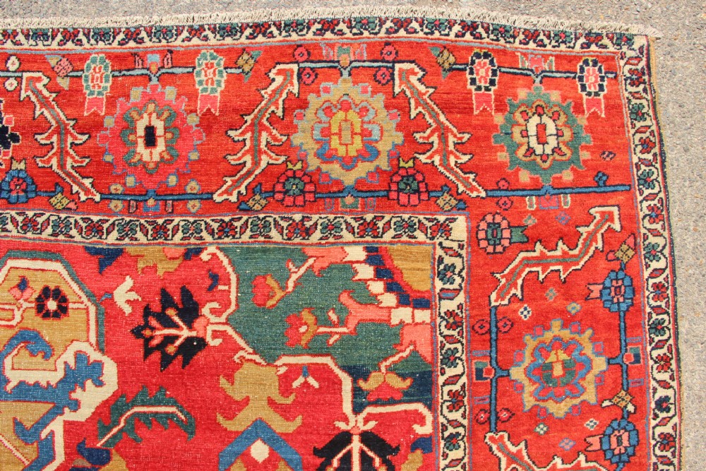 A large hand-knotted Persian Serapi carpet featuring a central geometric medallion in ochre and navy blue, surrounded by a navy field and wide terracotta red border. View IX