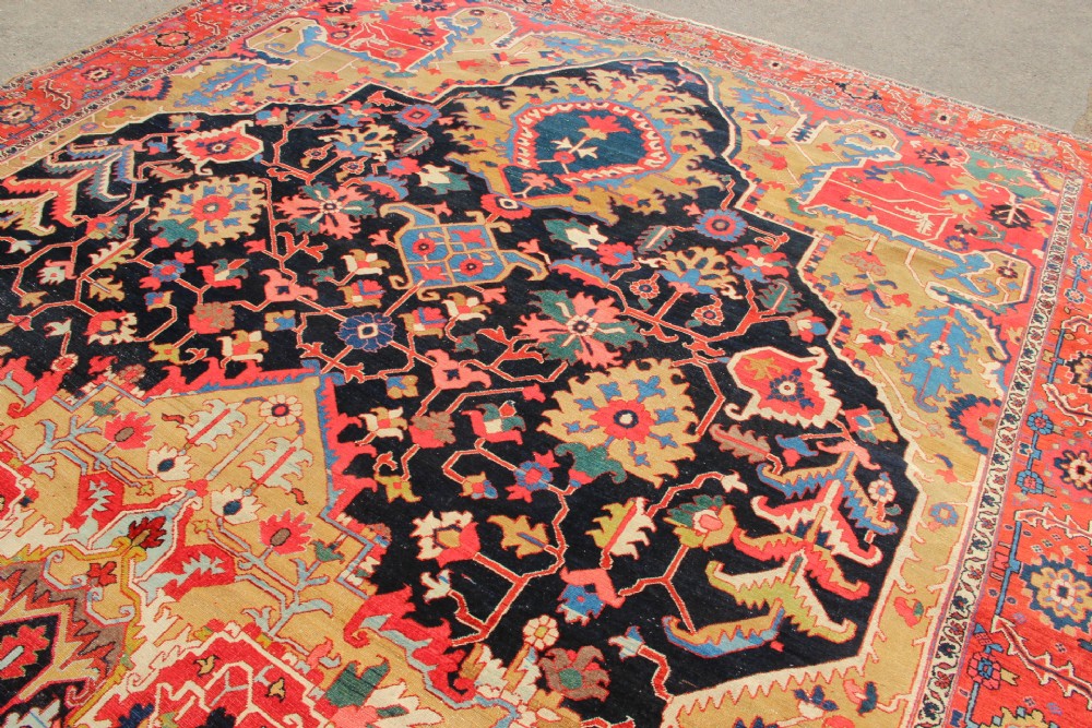 A large hand-knotted Persian Serapi carpet featuring a central geometric medallion in ochre and navy blue, surrounded by a navy field and wide terracotta red border. View X