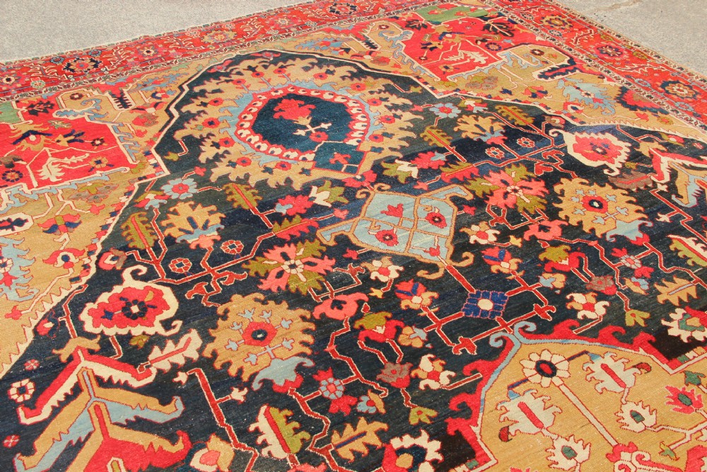 A large hand-knotted Persian Serapi carpet featuring a central geometric medallion in ochre and navy blue, surrounded by a navy field and wide terracotta red border. View XI