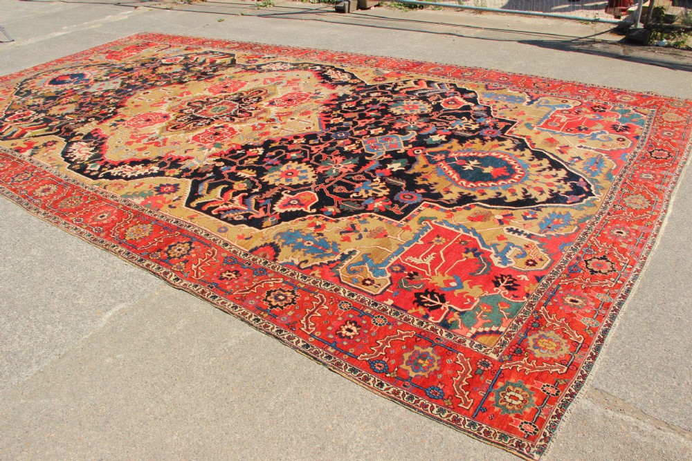 A large hand-knotted Persian Serapi carpet featuring a central geometric medallion in ochre and navy blue, surrounded by a navy field and wide terracotta red border. View XII