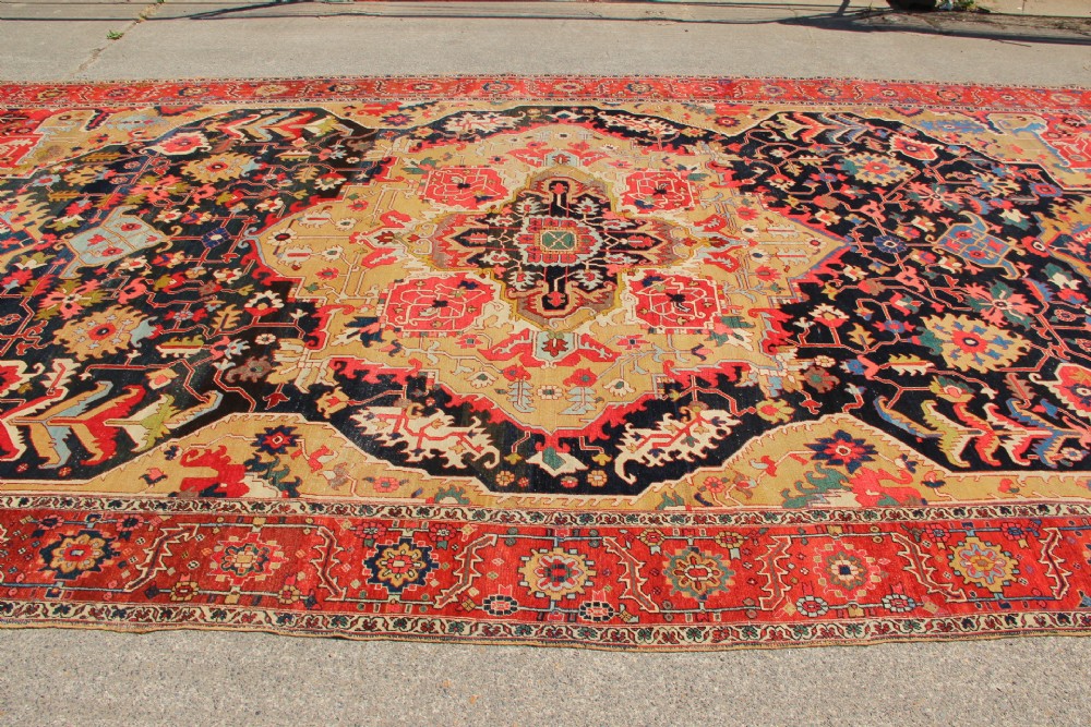 A large hand-knotted Persian Serapi carpet featuring a central geometric medallion in ochre and navy blue, surrounded by a navy field and wide terracotta red border. View XIII