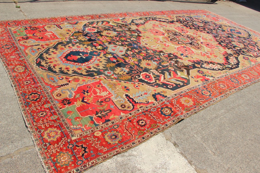 A large hand-knotted Persian Serapi carpet featuring a central geometric medallion in ochre and navy blue, surrounded by a navy field and wide terracotta red border. View XIV