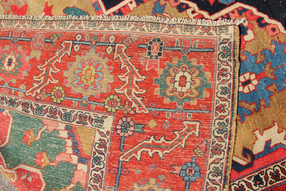 A large hand-knotted Persian Serapi carpet featuring a central geometric medallion in ochre and navy blue, surrounded by a navy field and wide terracotta red border. View XV