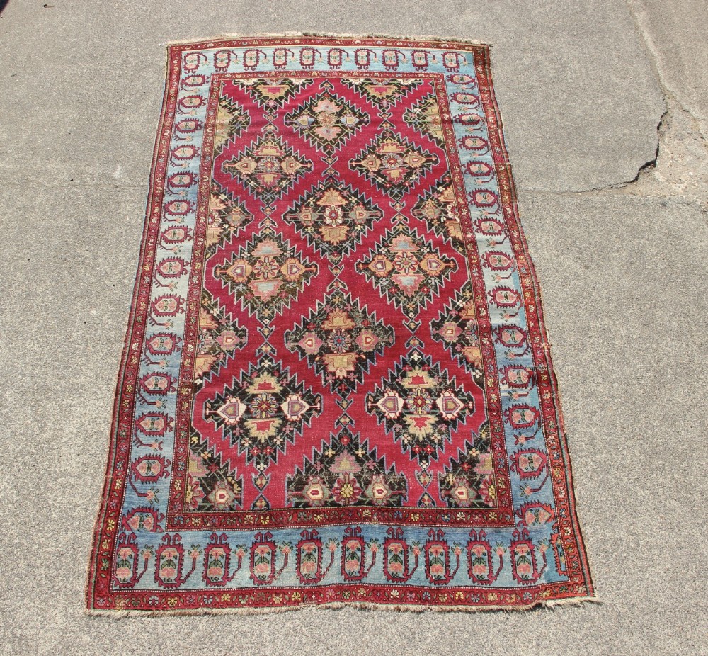 An antique Caucasian Karabagh carpet with a crimson red field displaying stepped geometric medallions framed by a sky blue border.