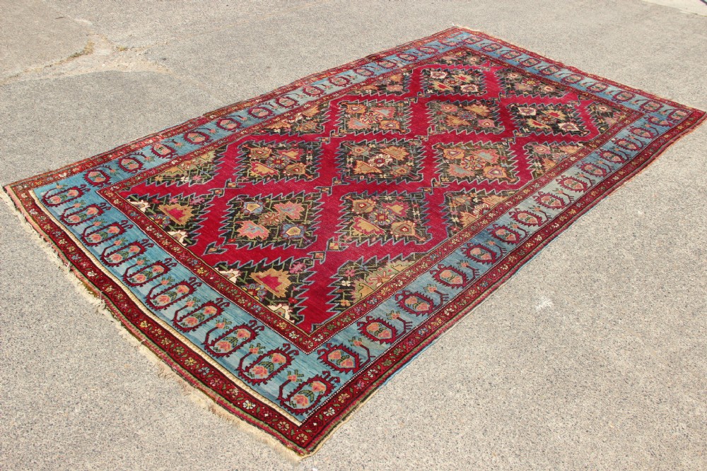 An antique Caucasian Karabagh carpet with a crimson red field displaying stepped geometric medallions framed by a sky blue border. View IV
