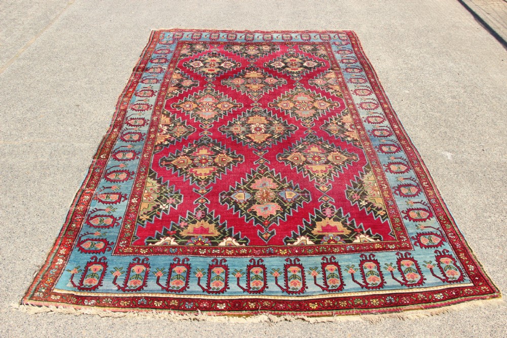 An antique Caucasian Karabagh carpet with a crimson red field displaying stepped geometric medallions framed by a sky blue border. View V
