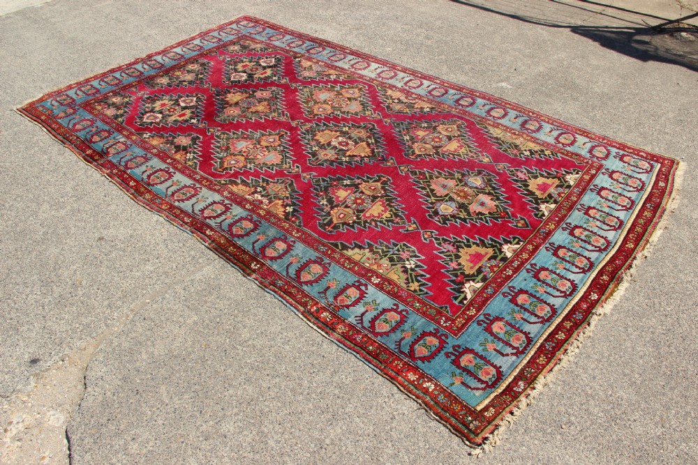 An antique Caucasian Karabagh carpet with a crimson red field displaying stepped geometric medallions framed by a sky blue border. View VI