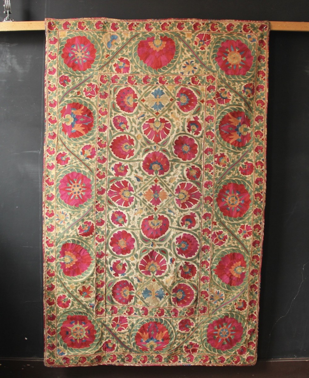 A rectangular hand-embroidered antique Suzani textile featuring red and pink floral medallions on a cream background with green vine borders.