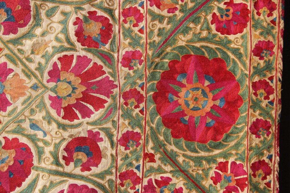 A rectangular hand-embroidered antique Suzani textile featuring red and pink floral medallions on a cream background with green vine borders. View V