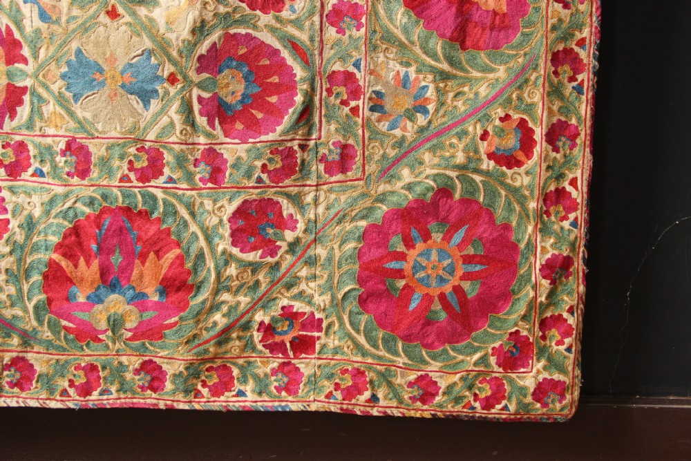 A rectangular hand-embroidered antique Suzani textile featuring red and pink floral medallions on a cream background with green vine borders. View VI