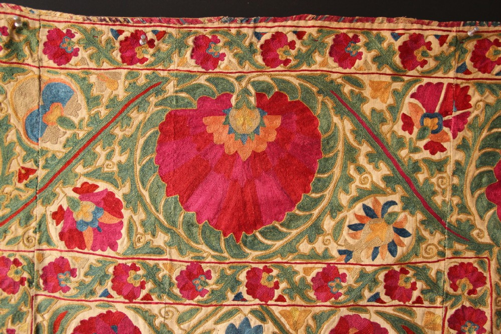 A rectangular hand-embroidered antique Suzani textile featuring red and pink floral medallions on a cream background with green vine borders. View X