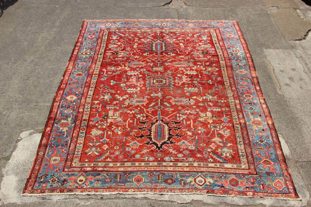An antique Persian Bakshaish wool carpet featuring a terracotta red field with geometric central motifs enclosed by a light blue patterned border.
