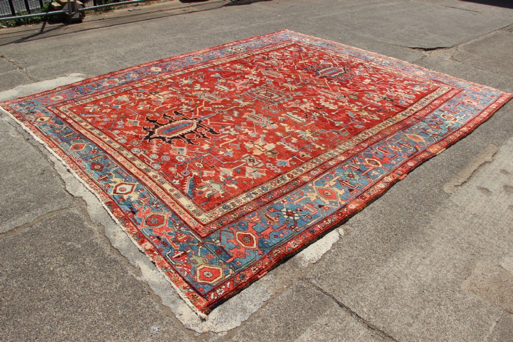 An antique Persian Bakshaish wool carpet featuring a terracotta red field with geometric central motifs enclosed by a light blue patterned border. View IV