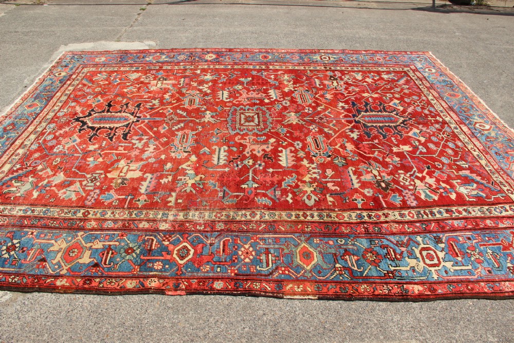 An antique Persian Bakshaish wool carpet featuring a terracotta red field with geometric central motifs enclosed by a light blue patterned border. View V