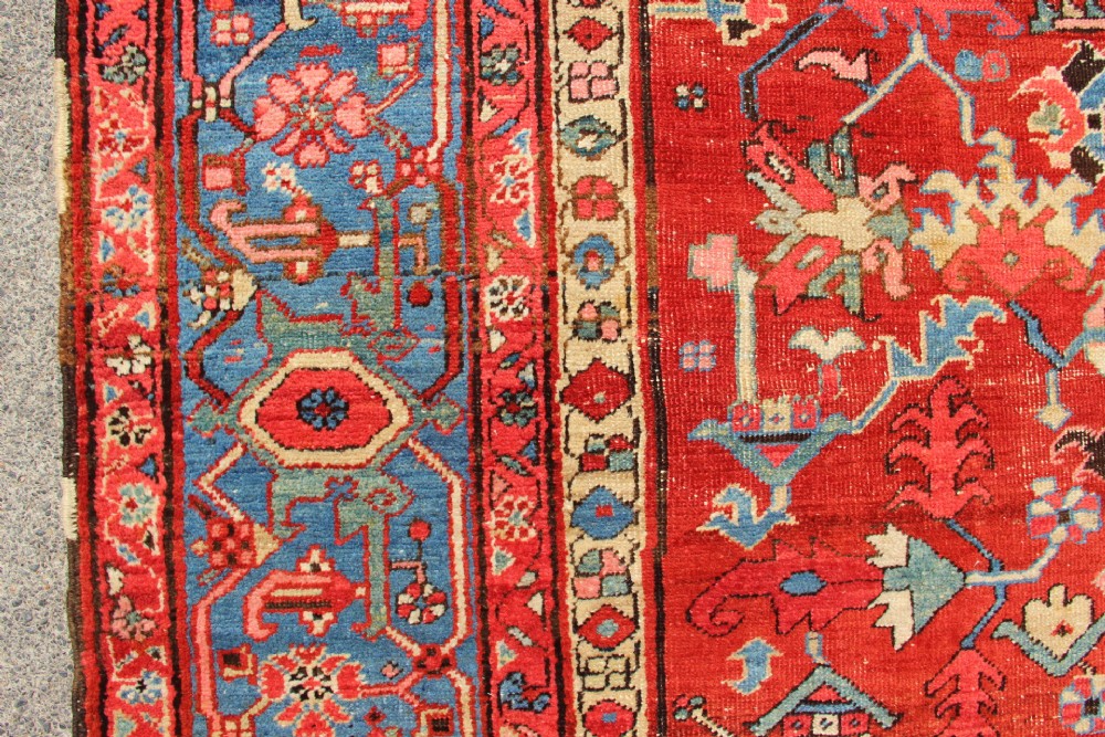 An antique Persian Bakshaish wool carpet featuring a terracotta red field with geometric central motifs enclosed by a light blue patterned border. View X