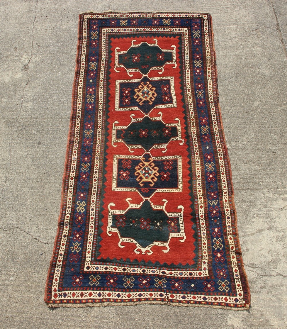 An antique Caucasian Kazak wool rug featuring central geometric medallions on a red ground enclosed by a navy blue patterned border.