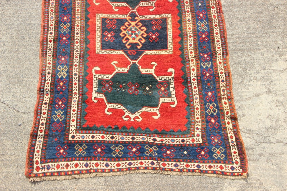 An antique Caucasian Kazak wool rug featuring central geometric medallions on a red ground enclosed by a navy blue patterned border. View II