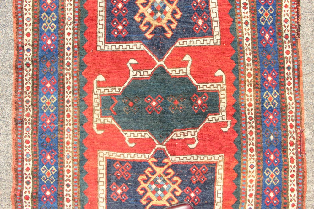 An antique Caucasian Kazak wool rug featuring central geometric medallions on a red ground enclosed by a navy blue patterned border. View III
