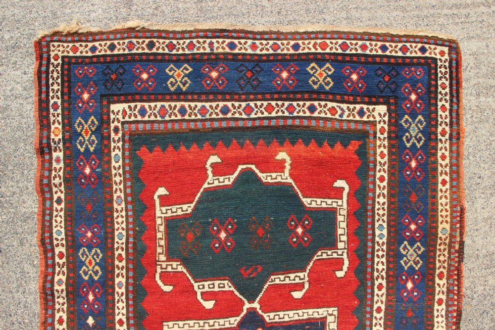 An antique Caucasian Kazak wool rug featuring central geometric medallions on a red ground enclosed by a navy blue patterned border. View IV