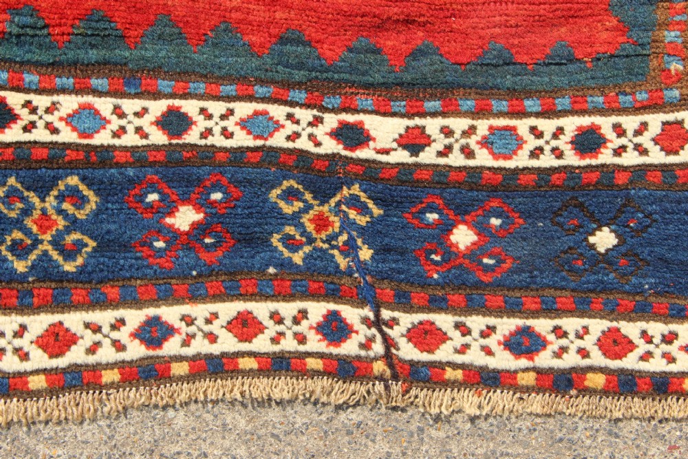 An antique Caucasian Kazak wool rug featuring central geometric medallions on a red ground enclosed by a navy blue patterned border. View V