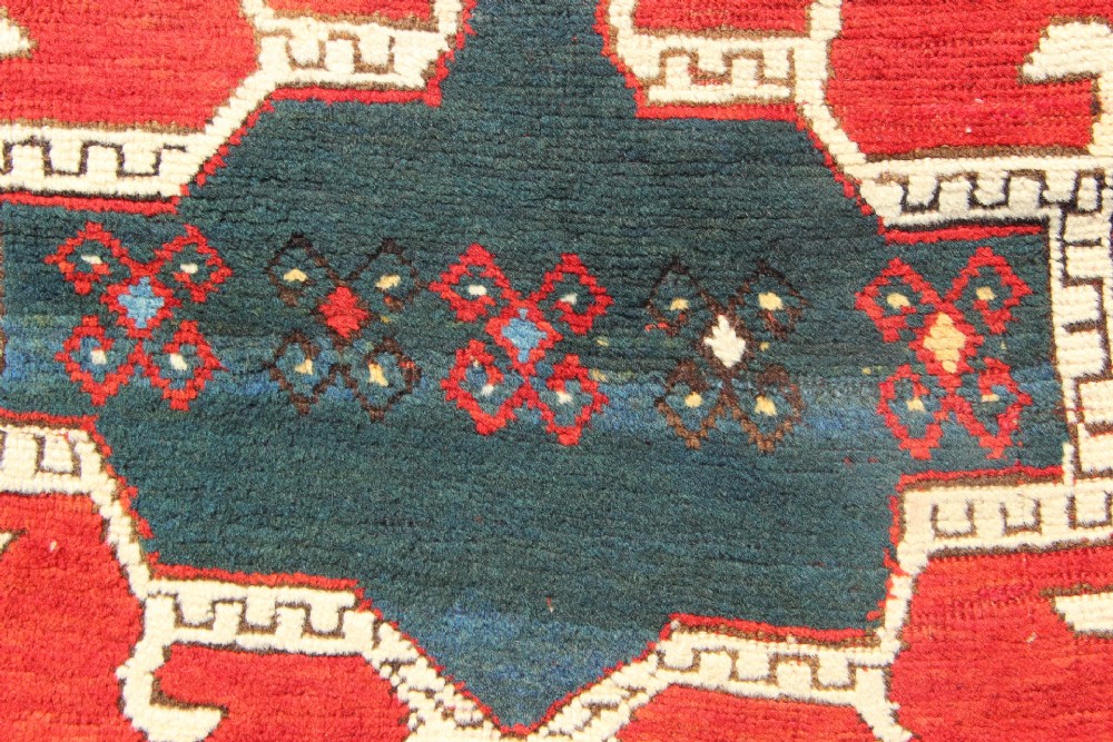 An antique Caucasian Kazak wool rug featuring central geometric medallions on a red ground enclosed by a navy blue patterned border. View VI