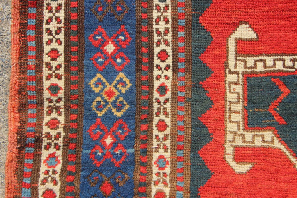 An antique Caucasian Kazak wool rug featuring central geometric medallions on a red ground enclosed by a navy blue patterned border. View VII