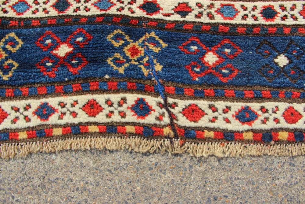 An antique Caucasian Kazak wool rug featuring central geometric medallions on a red ground enclosed by a navy blue patterned border. View VIII