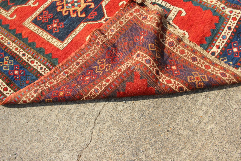 An antique Caucasian Kazak wool rug featuring central geometric medallions on a red ground enclosed by a navy blue patterned border. View IX