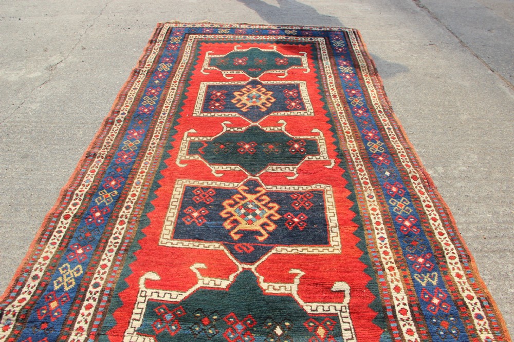An antique Caucasian Kazak wool rug featuring central geometric medallions on a red ground enclosed by a navy blue patterned border. View X