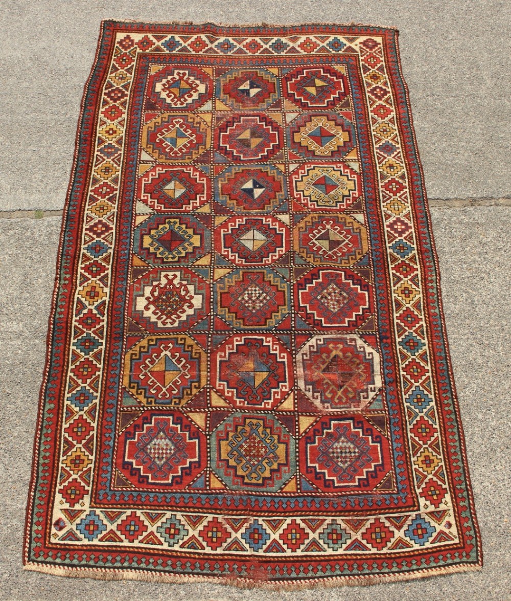 An antique Caucasian Moghan Kazak hand-knotted wool carpet displaying geometric octagonal medallions in red, blue, cream, and ochre yellow shades.