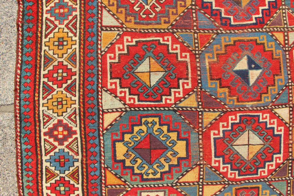 An antique Caucasian Moghan Kazak hand-knotted wool carpet displaying geometric octagonal medallions in red, blue, cream, and ochre yellow shades. View V