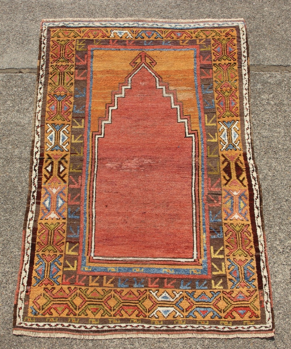A hand-knotted mid-19th-century Konya prayer rug featuring a central red stepped mihrab motif surrounded by geometric patterned borders in ochre yellow, brown, and blue tones.