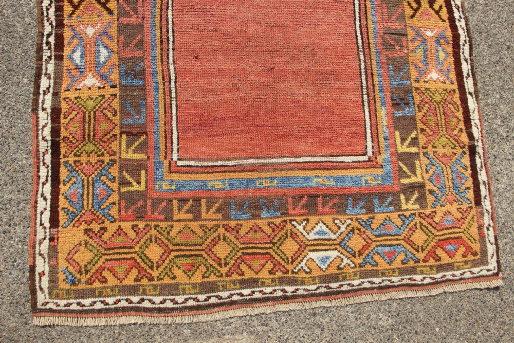 A hand-knotted mid-19th-century Konya prayer rug featuring a central red stepped mihrab motif surrounded by geometric patterned borders in ochre yellow, brown, and blue tones. View V