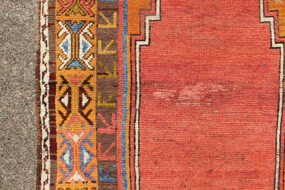 A hand-knotted mid-19th-century Konya prayer rug featuring a central red stepped mihrab motif surrounded by geometric patterned borders in ochre yellow, brown, and blue tones. View VI