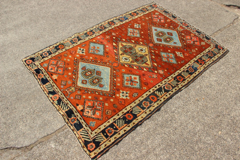 An antique hand-knotted Heriz rug featuring geometric diamond medallions in blue and yellow set against a rust-red ground with a patterned dark border. View II