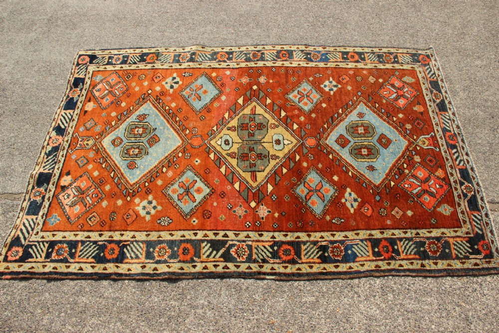An antique hand-knotted Heriz rug featuring geometric diamond medallions in blue and yellow set against a rust-red ground with a patterned dark border. View III