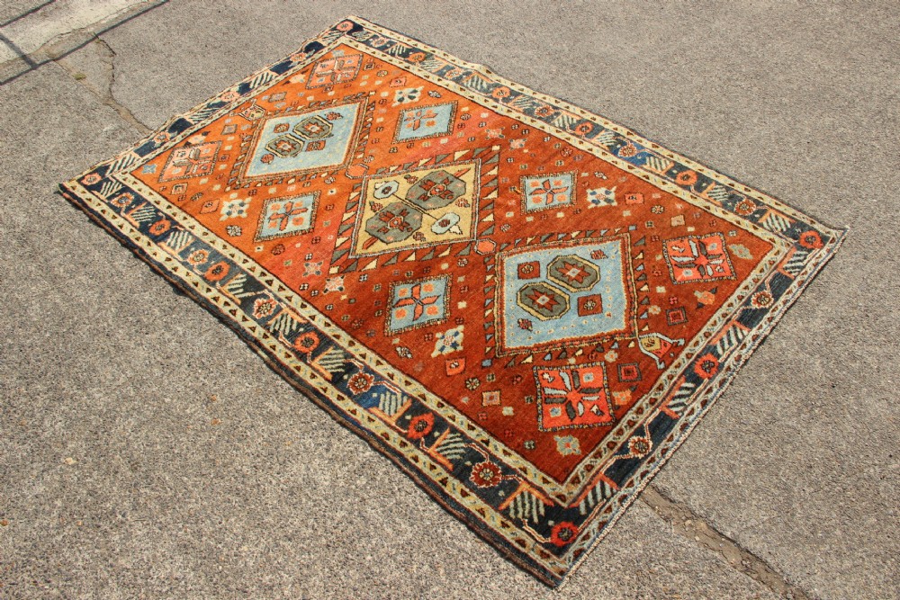 An antique hand-knotted Heriz rug featuring geometric diamond medallions in blue and yellow set against a rust-red ground with a patterned dark border. View IV