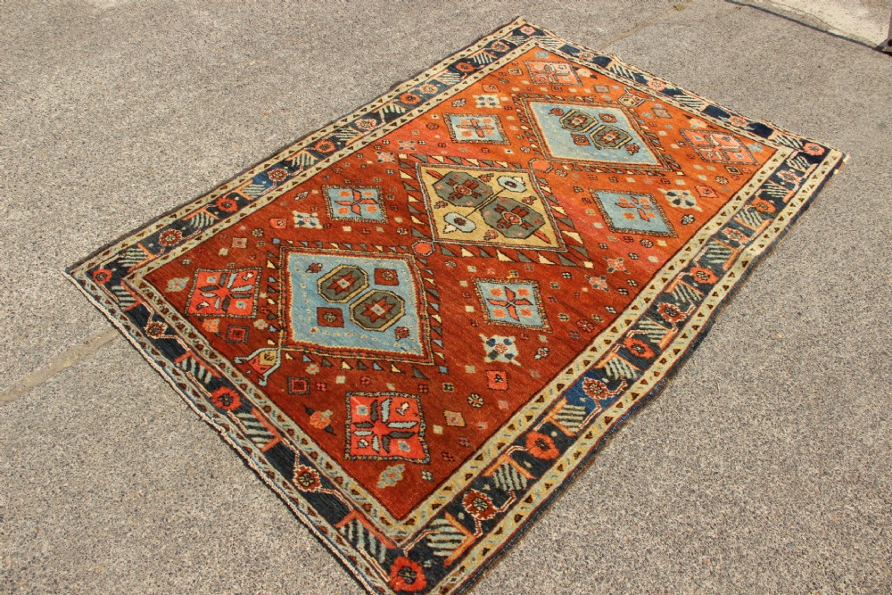 An antique hand-knotted Heriz rug featuring geometric diamond medallions in blue and yellow set against a rust-red ground with a patterned dark border. View V