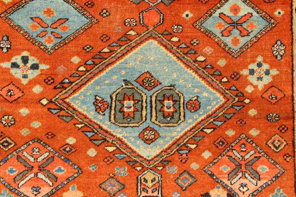 An antique hand-knotted Heriz rug featuring geometric diamond medallions in blue and yellow set against a rust-red ground with a patterned dark border. View VI