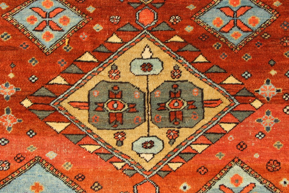 An antique hand-knotted Heriz rug featuring geometric diamond medallions in blue and yellow set against a rust-red ground with a patterned dark border. View VII