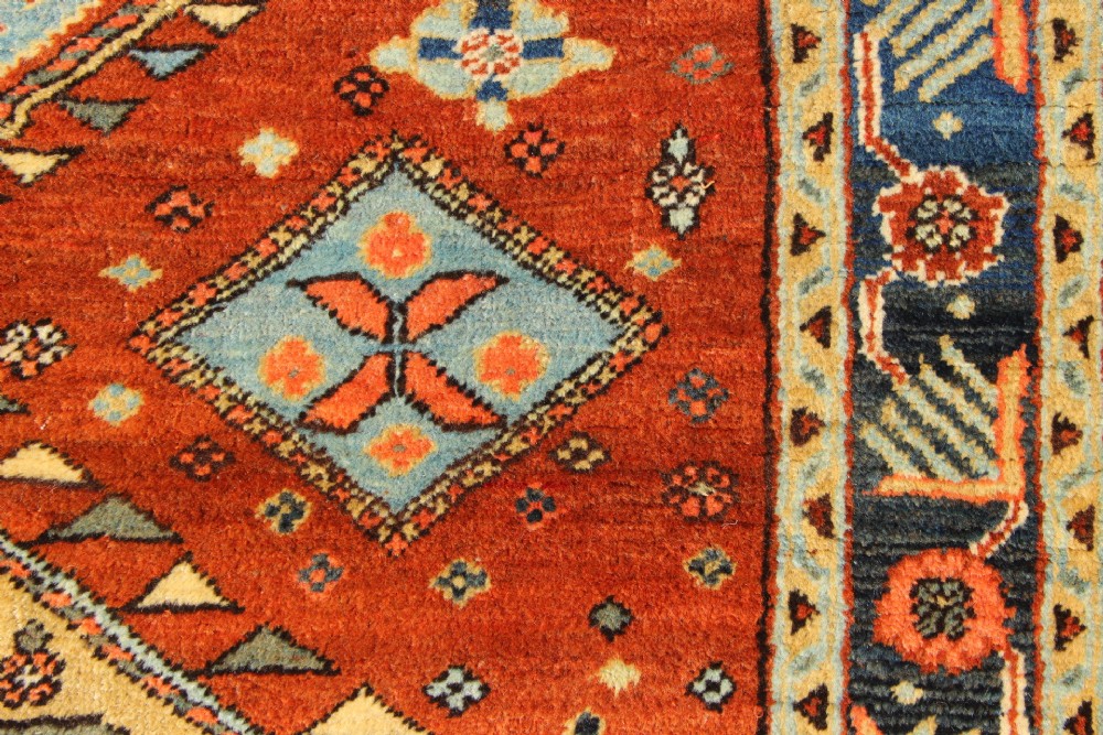 An antique hand-knotted Heriz rug featuring geometric diamond medallions in blue and yellow set against a rust-red ground with a patterned dark border. View VIII