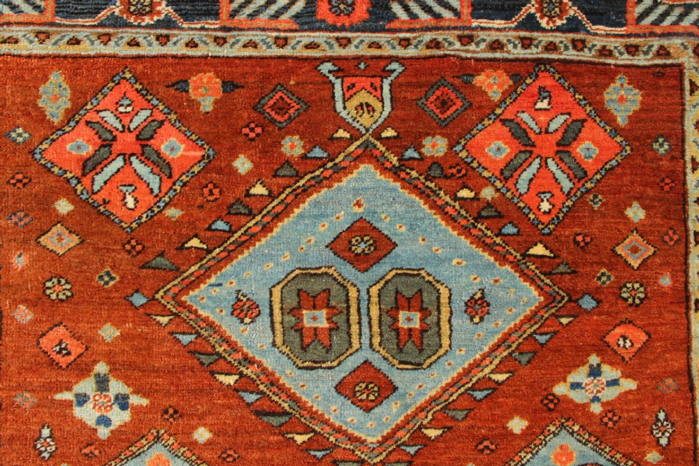 An antique hand-knotted Heriz rug featuring geometric diamond medallions in blue and yellow set against a rust-red ground with a patterned dark border. View IX