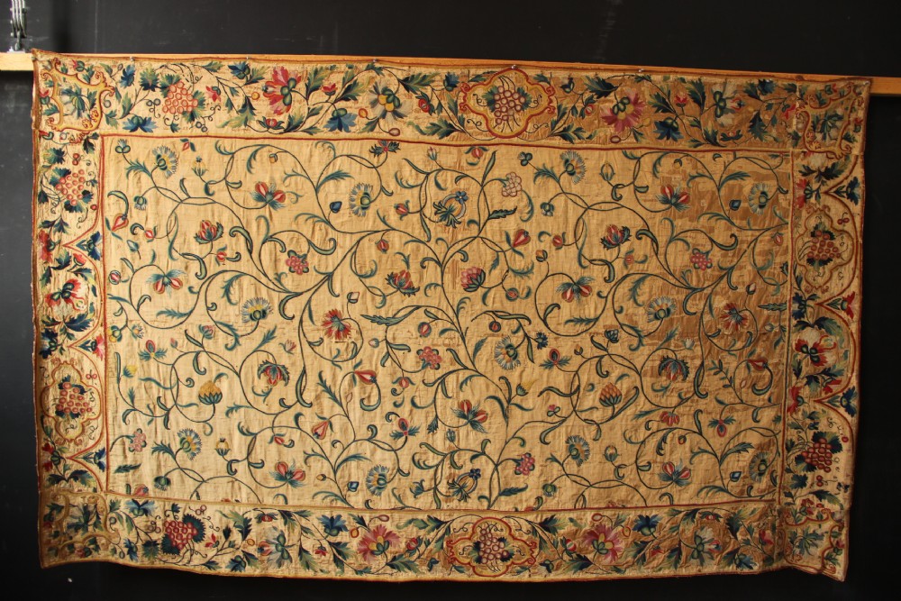 A large rectangular antique English crewelwork panel with intricate hand-embroidered floral tendrils in green, blue, and red wool on a cream background.