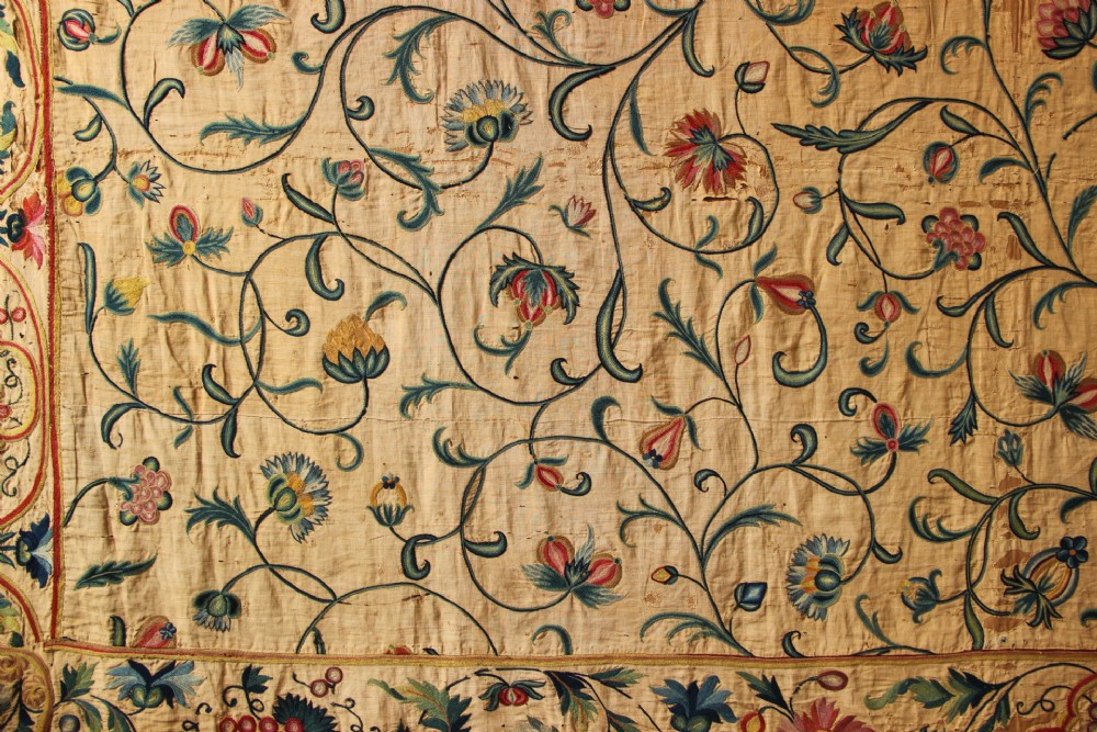 A large rectangular antique English crewelwork panel with intricate hand-embroidered floral tendrils in green, blue, and red wool on a cream background. View IV