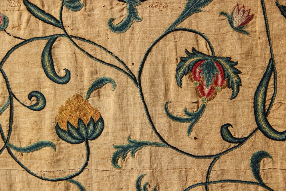 A large rectangular antique English crewelwork panel with intricate hand-embroidered floral tendrils in green, blue, and red wool on a cream background. View V