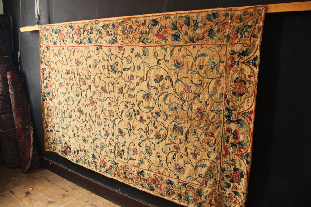A large rectangular antique English crewelwork panel with intricate hand-embroidered floral tendrils in green, blue, and red wool on a cream background. View XV