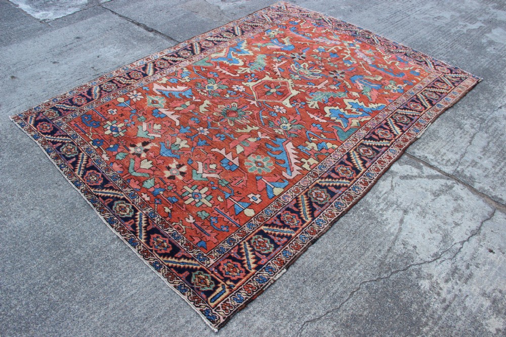 An antique hand-knotted Heriz carpet featuring a terracotta red field with central geometric motifs and a navy blue patterned border. View V