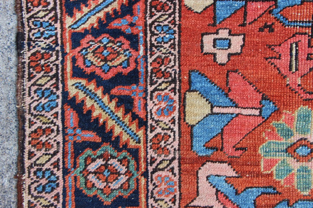 An antique hand-knotted Heriz carpet featuring a terracotta red field with central geometric motifs and a navy blue patterned border. View VIII